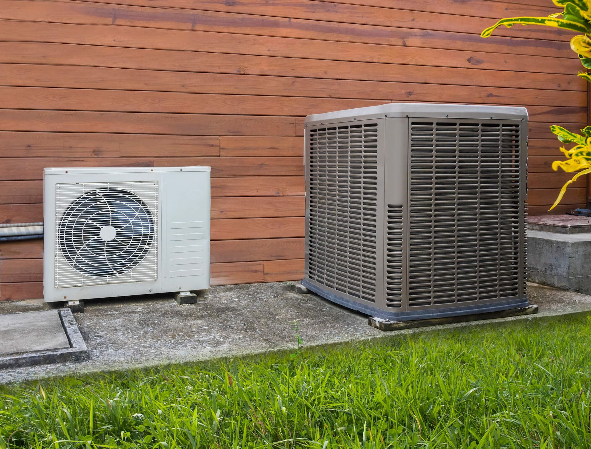 Two air conditioners are sitting next to each other on the side of a house.